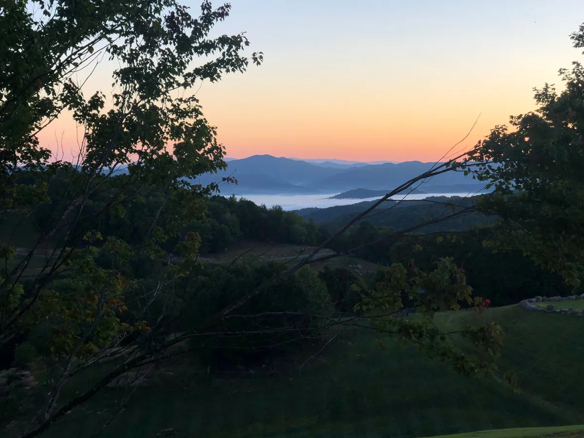 A panoramic view of mountains, a sparkling lake, and lush trees beneath a wide sky, evoking tranquility and connection with nature. The scene suggests opportunities for sunrise watching, peaceful strolls, and moments of reflection amidst stunning landscapes.