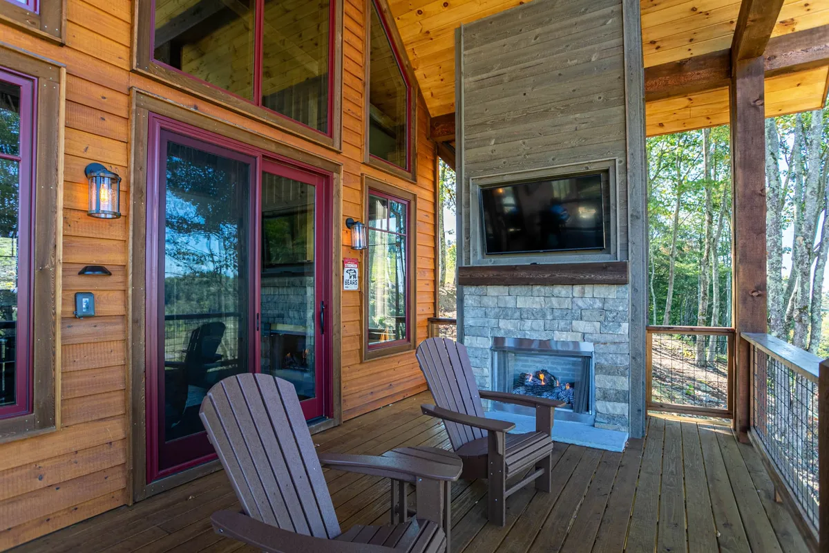 An inviting outdoor living space with Adirondack chairs circling a stone fireplace, large glass doors opening to the interior, wooden beams, and a view that evokes relaxation and social gatherings. The area suggests both daytime and evening enjoyment, perfect for winding down after an active day.