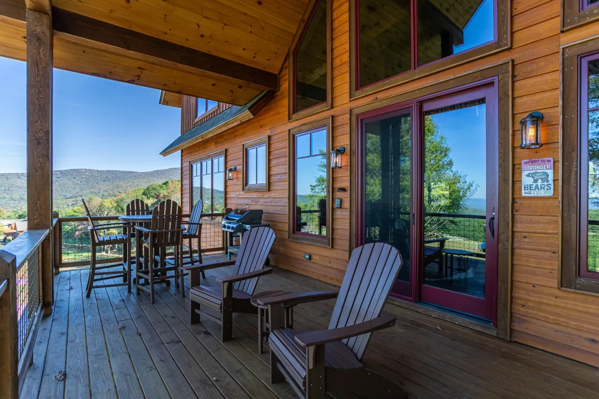 The photo features a spacious wooden deck with outdoor seating and a grill, all overlooking a scenic mountain vista. The area is perfect for socializing, relaxing, and enjoying meals outside while taking in the beautiful surroundings.
