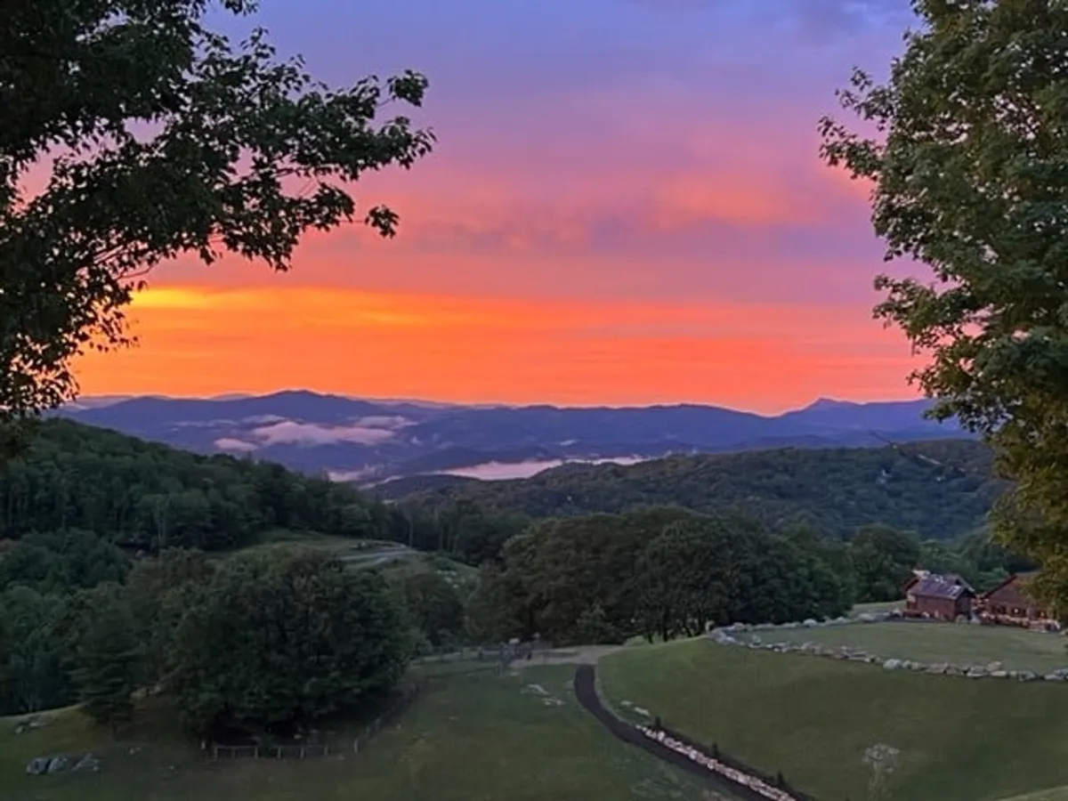 A breathtaking view of a sunset over a mountain range, with a meandering path and abundant greenery leading to a distant cabin. The image conveys tranquility, natural beauty, and the sense of exploration and outdoor enjoyment available at Eagles Nest.
