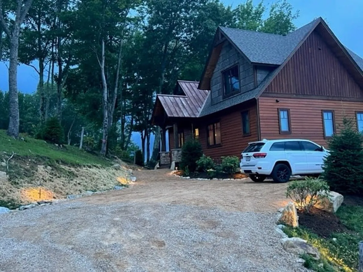 The exterior of Evergreen with a cozy wooden house, a gravel driveway, and thick forest surroundings. The property’s inviting porch, ample outdoor space, and natural setting emphasize a peaceful, dog-friendly environment for both play and relaxation.