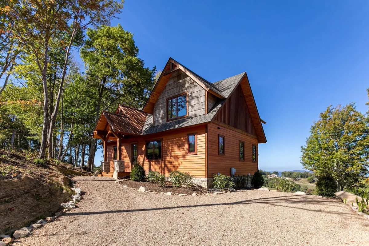 This exterior image showcases the wooden facade of Evergreen, featuring large windows and stone accents, surrounded by mature trees. The inviting porch and natural setting are visible, creating a welcoming atmosphere ideal for pet owners looking for outdoor relaxation and easy access to nature.