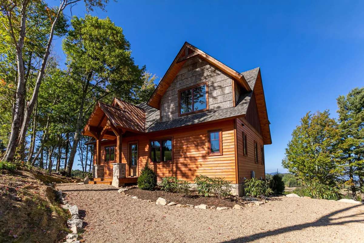 The image showcases the exterior of a beautiful single-family mountain home with a warm wooden facade, expansive windows that capture the scenery, and a sturdy stone foundation. The landscaped garden frames a welcoming porch, perfect for relaxing while soaking in sweeping mountain views. The scene is set at sunset, casting a golden glow that enhances the serene and inviting atmosphere.