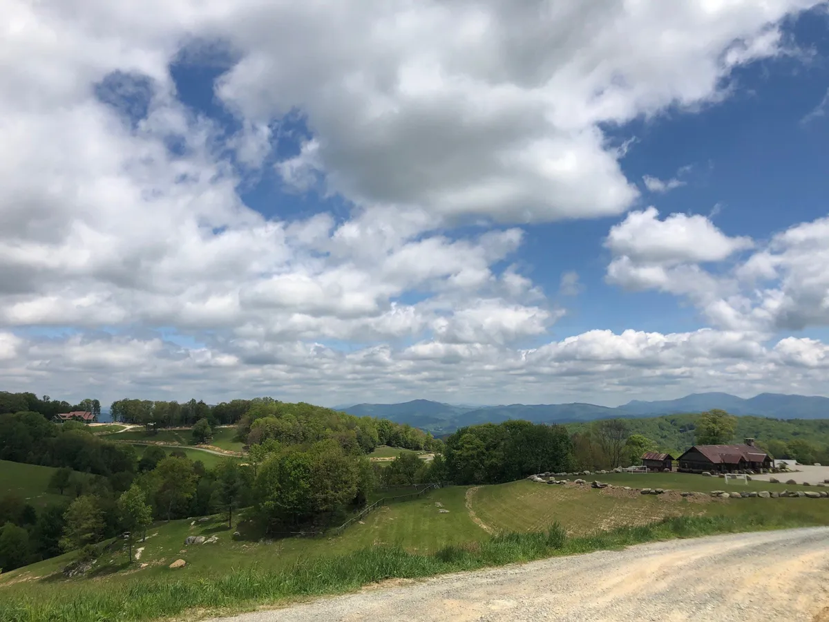 A sweeping panoramic view of rolling hills and distant mountains under a sky scattered with clouds. The foreground features open fields and subtle farm buildings, evoking a sense of serenity and natural beauty. There are no visible people, but the scene suggests ample space for hiking, picnicking, and outdoor exploration.