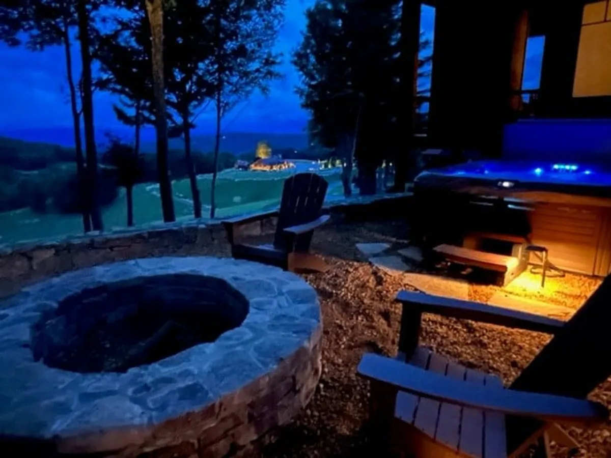 This exterior shot captures both the inviting stone fire pit surrounded by Adirondack chairs and the nearby bubbling hot tub, set against a backdrop of rolling hills and distant mountain views. The outdoor area looks perfect for evening gatherings, stargazing, and soaking in the natural serenity.