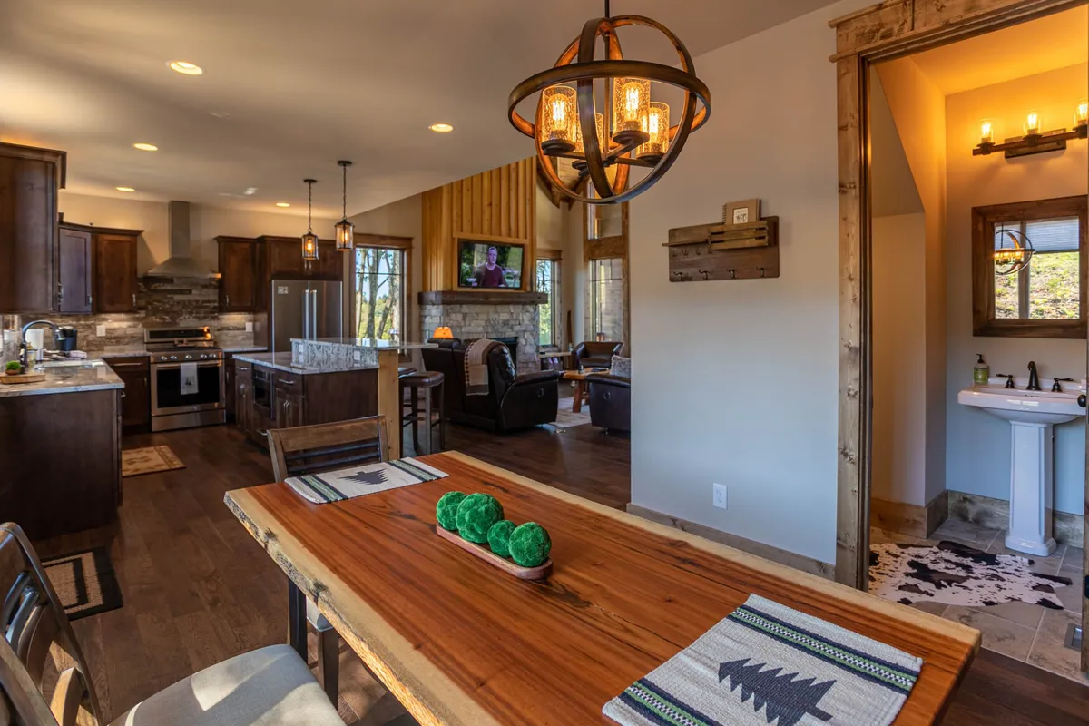 An inviting interior shot showing the dining table at the center, a kitchen island, a cozy living area with a fireplace, and decorative lighting. The space is set up for family meals, game nights, and relaxing by the fire, showcasing the open flow between kitchen, dining, and living spaces.