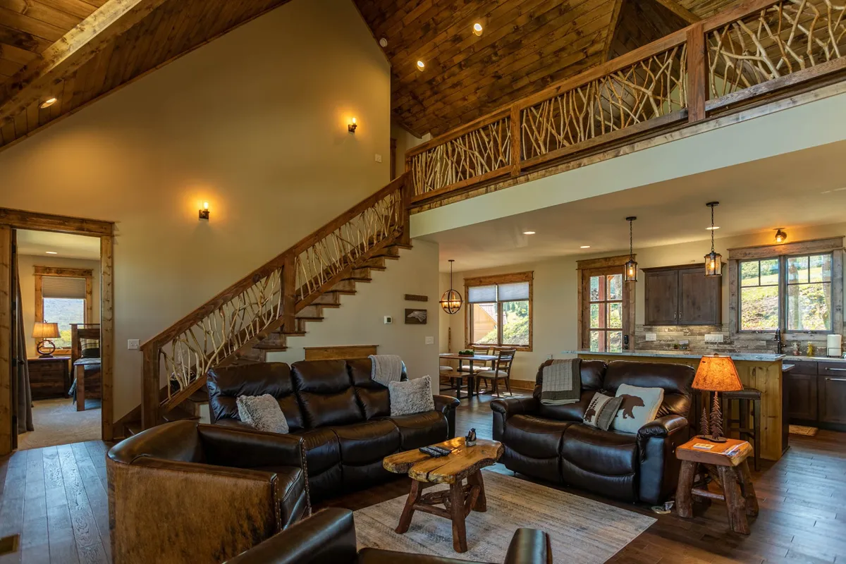A spacious open-concept living area featuring plush leather sofas, a wooden coffee table, large windows that let in abundant natural light, a visible kitchen area, and a wooden staircase. The space exudes warmth and invites gatherings, with the living room flowing seamlessly into the kitchen and dining area.