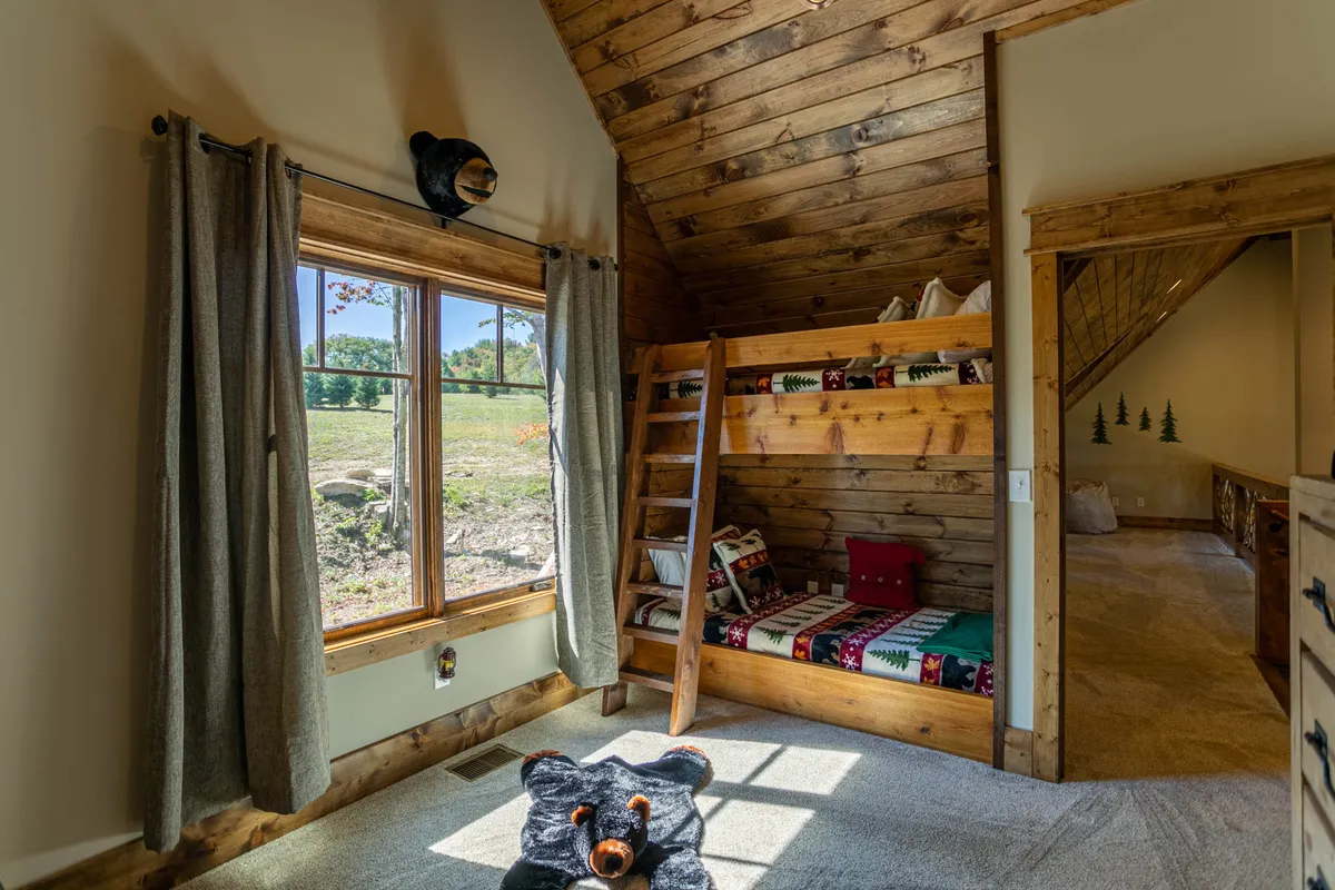 A playful and inviting bunk bedroom featuring built-in bunk beds, a large window with views of the outdoors, a whimsical bear rug, and warm wood paneling. Ideal for kids or friends, the room feels both fun and cozy.