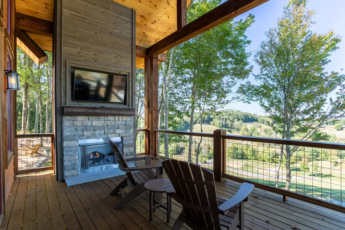 This inviting deck scene includes an outdoor fireplace, deck chairs, a mounted TV, and beautiful views of the trees and landscape beyond. The space is ideal for watching the game, cozying up by the fire, or simply enjoying the outdoors in comfort.