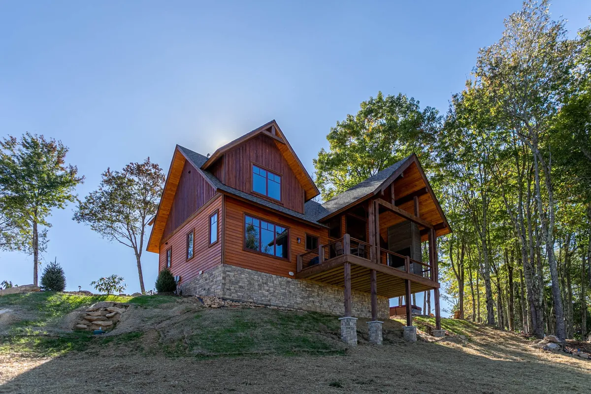 The exterior of a welcoming mountain home framed by large windows, a stone foundation, and a balcony overlooking the surrounding forest. Tall trees envelop the property, and the balcony hints at direct engagement with nature.